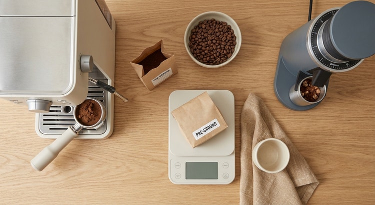 Flat lay of a home espresso workspace showing common mistakes — a kitchen scale without tare, a portafilter with uneven coffee distribution, a bag of pre-ground coffee next to whole beans, and a grinder with visible clumps in the chute, warm overhead light casting minimal shadows on a light wood surface