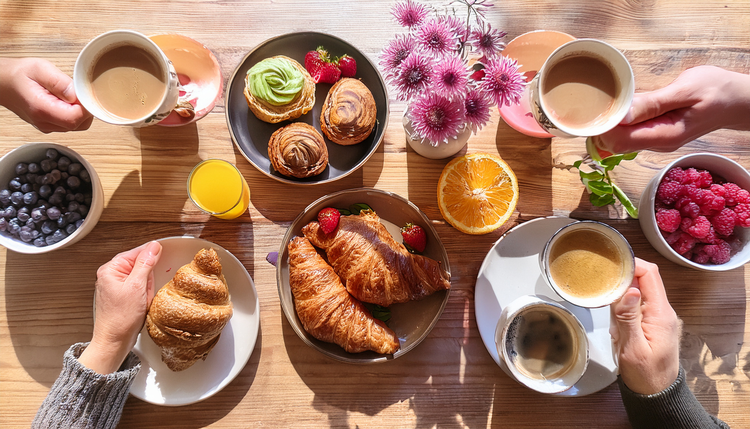 A sunlit brunch table with pastries, fresh fruit, and avocado toast, four different coffee drinks visible, friends' hands reaching across the table