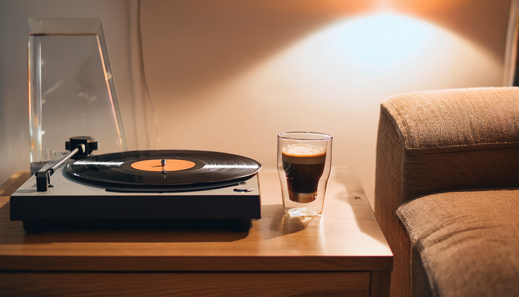 A turntable playing a vinyl record beside an espresso in a double-wall glass on a credenza, album covers leaning against the wall, warm lamp light