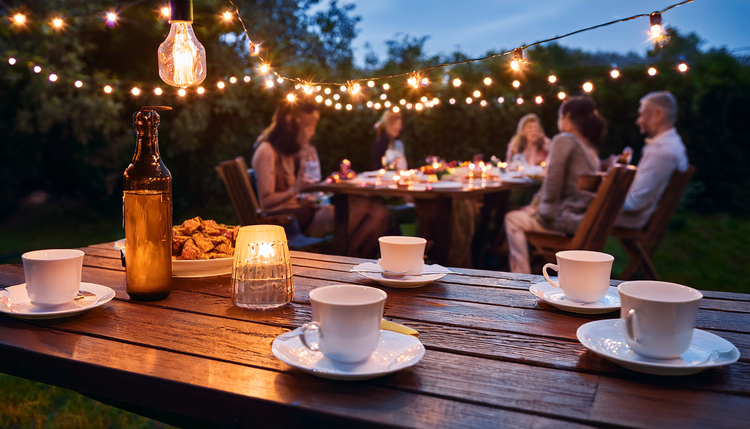A garden table under string lights at dusk with espresso cups among scattered dessert plates, friends around a barbecue in the background