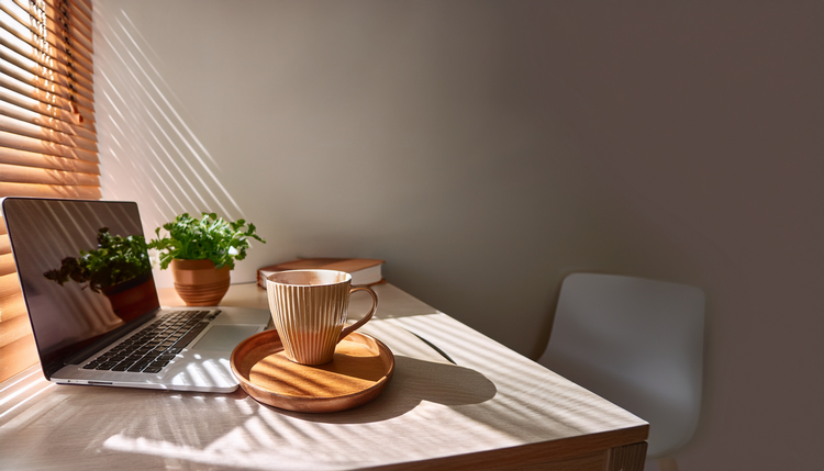 A minimalist home office desk with a laptop, notebook, and a freshly made cappuccino in a ceramic cup on a small copper tray, midmorning light through wooden blinds