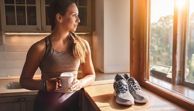 A runner leaning against a kitchen counter in running shoes and athletic wear, drinking espresso from a small ceramic cup, a smartwatch visible on their wrist