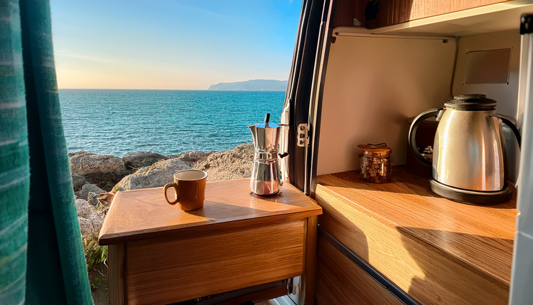 A compact portable espresso maker on a fold-out wooden shelf inside the open side door of a converted camper van, Mediterranean coastline visible through the doorframe