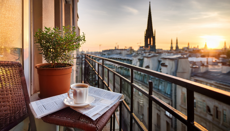A small city apartment balcony with a single espresso cup on the railing, rooftops and church spires visible in soft morning light