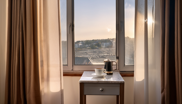 A boutique hotel room with a small espresso machine on a marble console, a freshly made espresso beside French doors opening onto a balcony with a city rooftop view