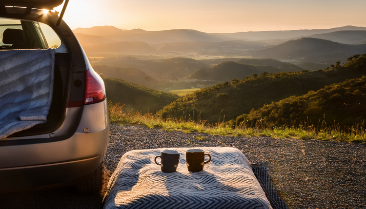 A portable espresso setup on the tailgate of a car at a scenic overlook, two cups ready, a winding road disappearing into rolling hills behind