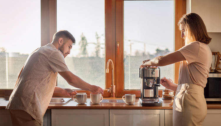 Two people in a bright modern kitchen, one operating an espresso machine with copper accents while the other reads a tablet at the counter, two ceramic cups ready