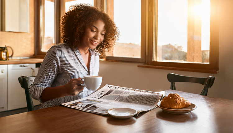 A person at a wooden kitchen table on a lazy Sunday morning with a broadsheet newspaper spread wide, a half-finished cappuccino with latte art in a wide ceramic cup