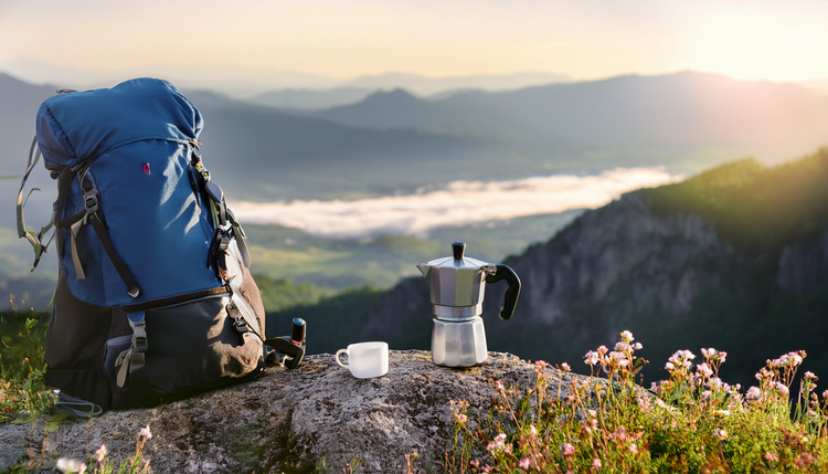 A portable hand-powered espresso maker and a small cup on a flat rock at a mountain overlook, a hiking backpack leaning against the rock, panoramic valley view