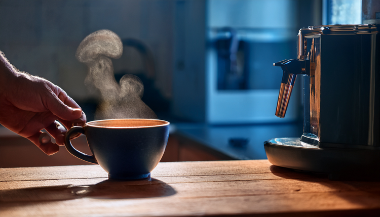 A kitchen in pre-dawn blue light with a single espresso cup steaming on a wooden counter, the only warm light from an espresso machine's copper-accented power indicator