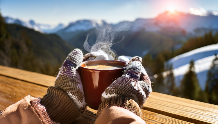Hands in fingerless gloves wrapping around a steaming espresso cup on a rustic wooden table at a mountain lodge terrace, snow-covered peaks in the background