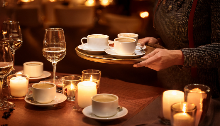 A candlelit dining table with dessert plates and wine glasses, a host carrying a tray of small white espresso cups from the kitchen