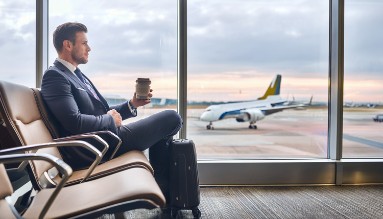A business traveler at an airport window seat holding an espresso cup while watching aircraft on the tarmac through floor-to-ceiling windows