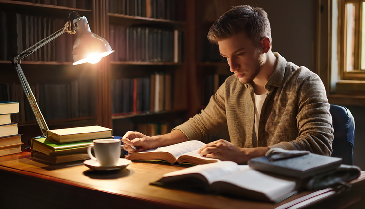 A university student studying at a wooden desk with open textbooks, highlighted notes, and a small espresso cup on a ceramic saucer, laptop open beside the books