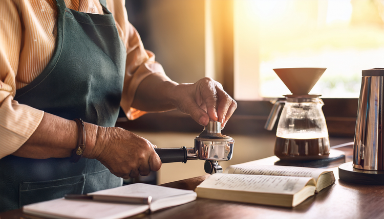 An older person's hands carefully tamping espresso grounds into a portafilter at a sunlit kitchen counter, reading glasses beside an open espresso guidebook
