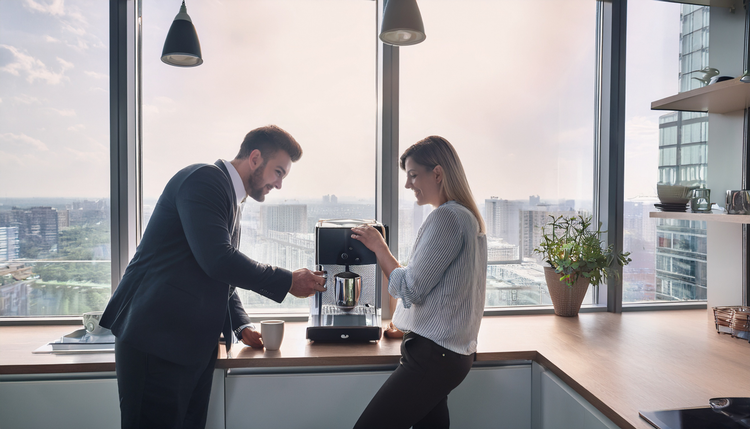 A modern bright office kitchen with two colleagues at a counter, one operating a matte black espresso machine while the other laughs mid-conversation