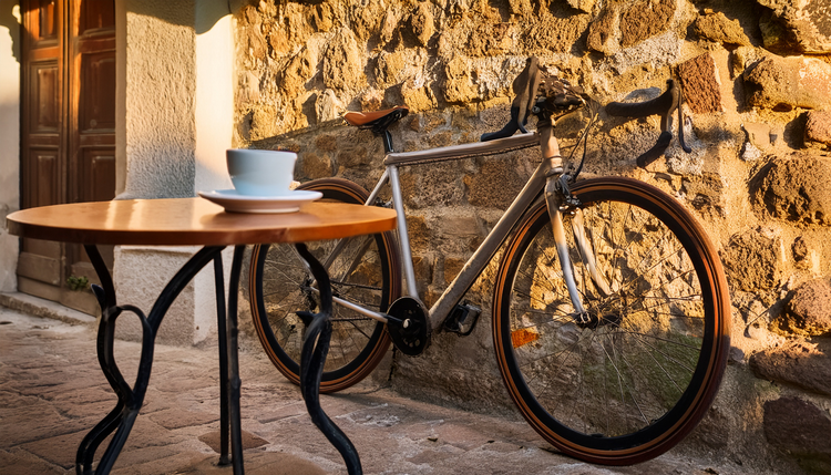 A road bicycle leaning against a sunlit stone wall beside a small cafe table with a double espresso in a ceramic cup, cycling gloves and sunglasses on the table