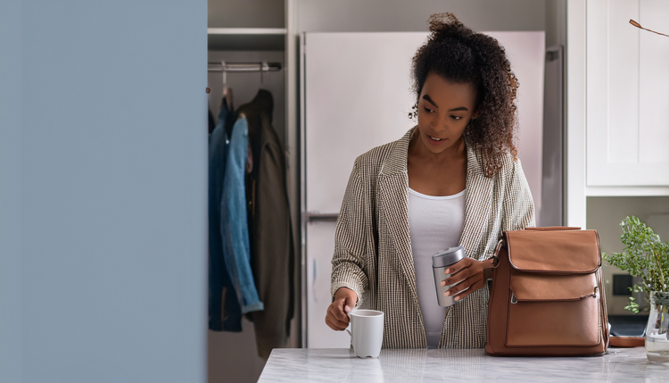 A person grabbing a stainless steel travel mug of espresso from a marble kitchen counter while picking up keys and a messenger bag, early morning grey-blue light through a hallway window