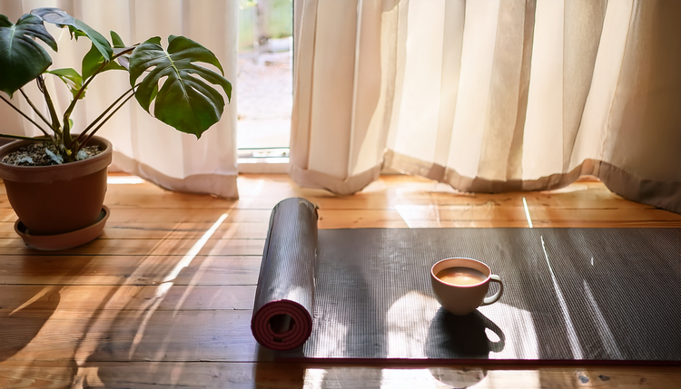 A rolled yoga mat on a wooden floor beside a low wooden bench holding a ceramic cup of espresso, soft morning light through sheer curtains
