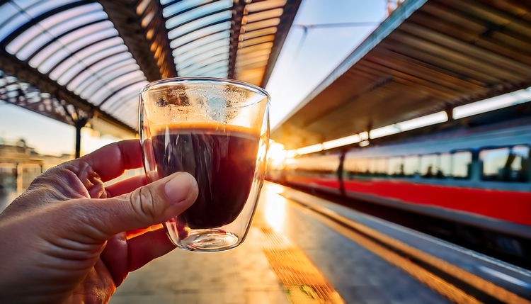 A hand holding a double-wall glass cup of espresso on a modern train station platform at dawn, blurred commuter train arriving in the background