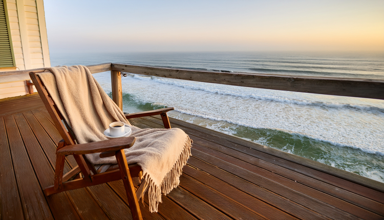 A weathered wooden deck of a beach house with a ceramic cup of espresso on the railing, the ocean visible in soft morning haze