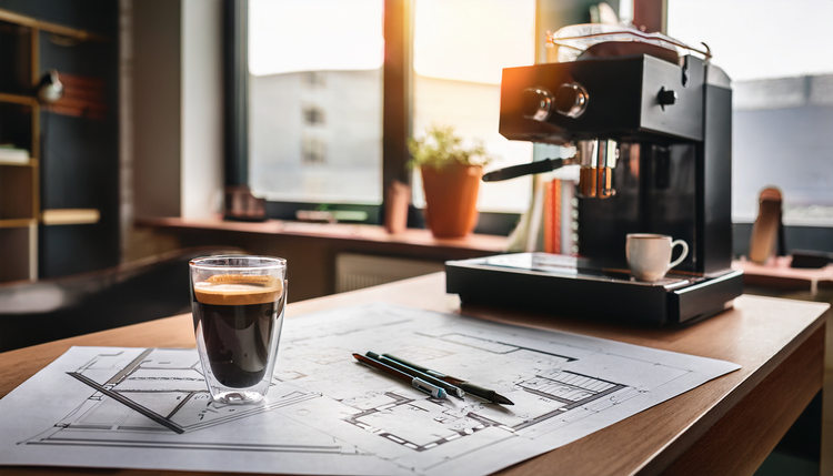 An architect's studio table with blueprints, material swatches, and a double espresso in a clear glass beside a copper mechanical pencil